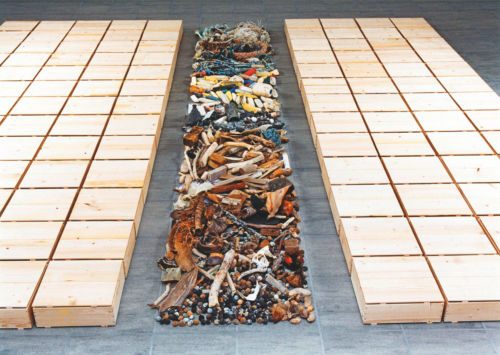 Bodeninstallation: ein langer Streifen aus Schwemmholz, Steinen und Strandgut zwischen zwei Flächen aus Holzkisten