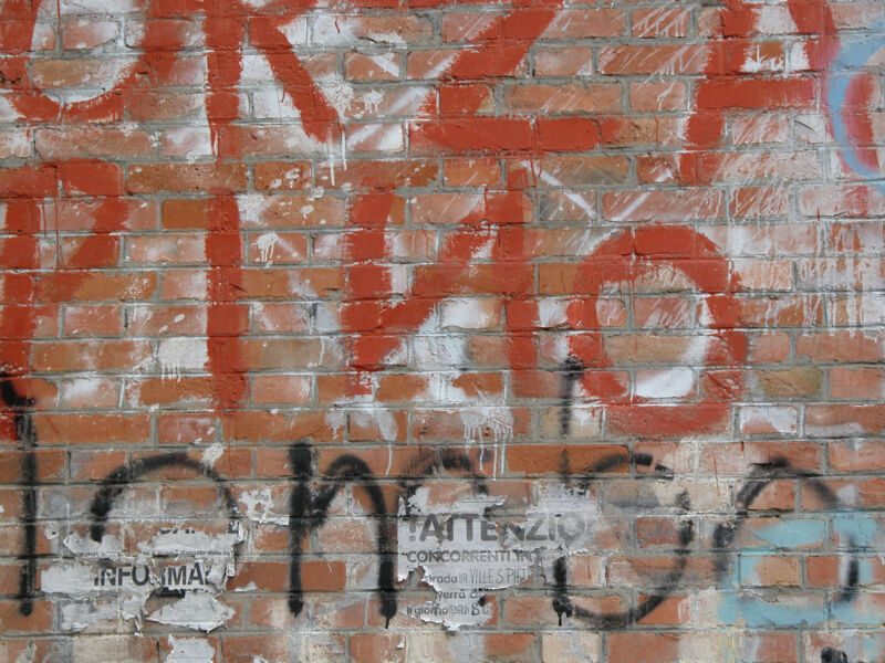 Backsteinmauer mit grossen roten und schwarzen Graffitibuchstaben und abgerissenen Plakatfetzen, Fotografie aus der Serie «Alltägliche Fundstücke I»