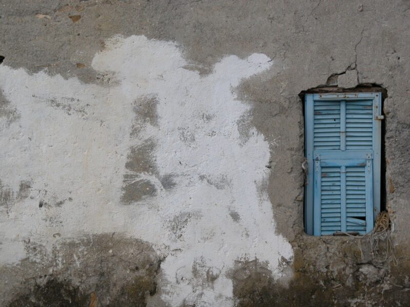 Graue, rissige Hausmauer mit grossem weissem Farbfleck und kleinem hellblauem Fensterladen, Fotografie aus der Serie «Alltägliche Fundstücke I»