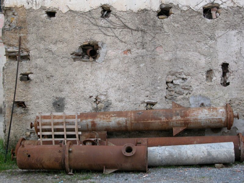 Verrostete Metallrohre und ein Gitter liegen vor einer alten Bruchsteinmauer mit Mauerlöchern, Fotografie aus der Serie «Alltägliche Fundstücke I»