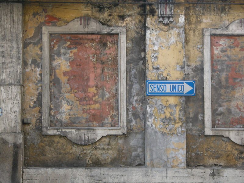 Verwitterte gelb-rote Fassade mit zwei leeren Zierrahmen und blauem Einbahnschild «Senso unico», Fotografie aus der Serie «Alltägliche Fundstücke I»