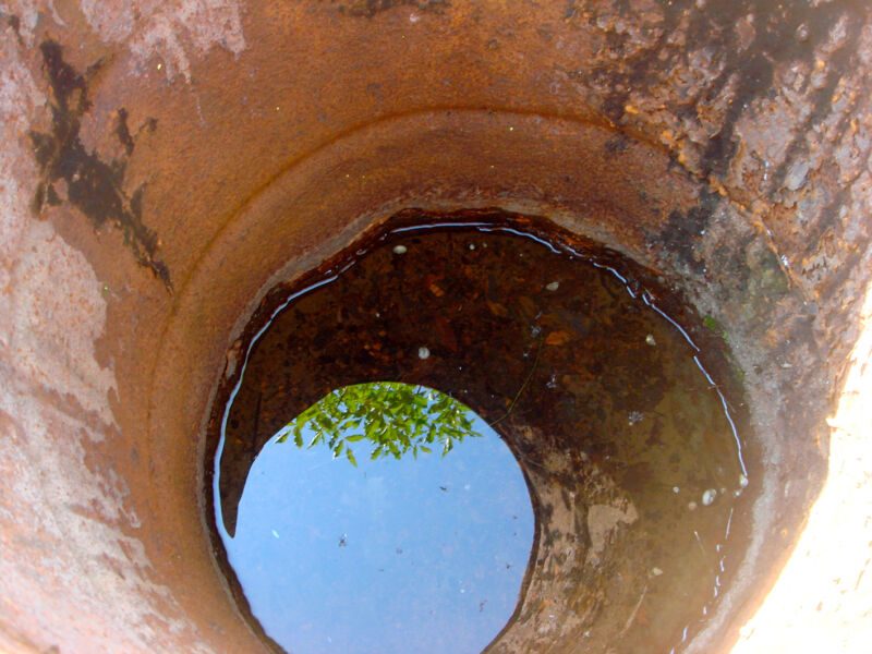 Blick in ein rostiges Rohr, in dessen Wasserspiegel blauer Himmel und grünes Laub erscheinen, Fotografie aus der Serie «Alltägliche Fundstücke I»