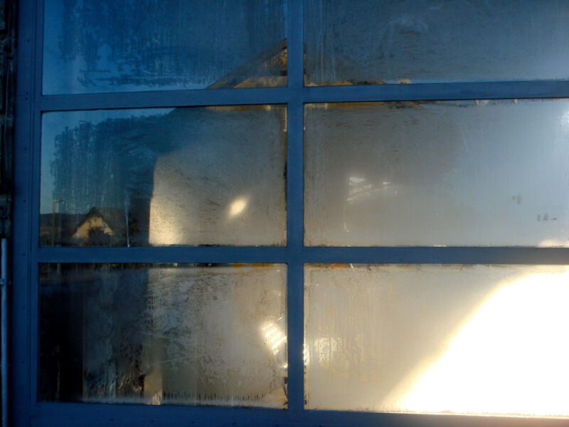 Beschlagenes Sprossenfenster im Gegenlicht mit hellem Lichtfleck und schemenhaft gespiegeltem Haus, Fotografie aus der Serie «Alltägliche Fundstücke I»