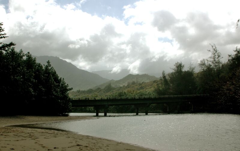 Flache Brücke über eine breite Flussmündung mit Sandufer, dahinter bewaldete Berge unter grauen Wolken, Fotografie aus der Serie «Hawaii»