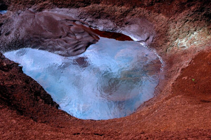 Kleine Wasserlache in roter Felsmulde, in der sich hellblau der Himmel spiegelt, Fotografie aus der Serie «Hawaii»