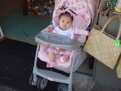 Kleinkind mit rosa Haarschleifen im Buggy neben geflochtenen Strohtaschen vor einem Laden, Fotografie aus der Serie «Honolulu»
