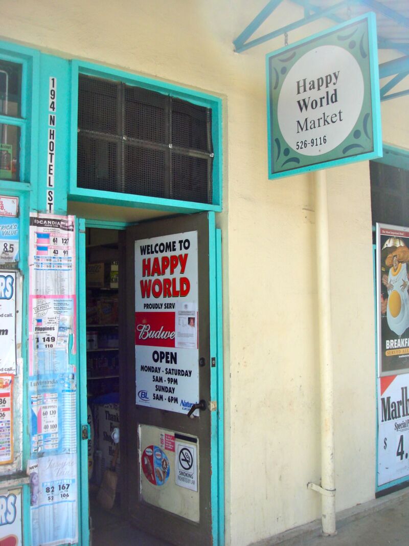 Türkis gerahmter Ladeneingang mit Schild «Happy World Market» und Plakaten an der hellen Fassade, Fotografie aus der Serie «Honolulu»