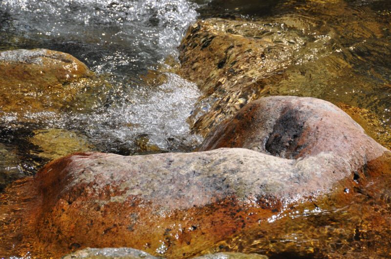 Rötlich-ockerfarbener, rund geschliffener Stein, von schäumendem Wasser umspült, Fotografie aus der Serie «An ligurischen Flüssen und Bächen»