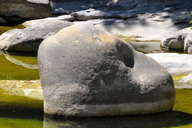 Grosser, rundlicher Granitblock steht in seichtem grünem Wasser, dahinter helle Felsen, Fotografie aus der Serie «An ligurischen Flüssen und Bächen»