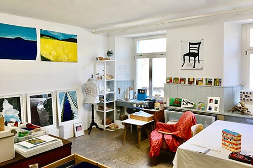 Helles Atelier von Katharina Rapp mit Bildern an den Wänden, Regal, Schneiderpuppe, rotem Sessel und Arbeitstisch
